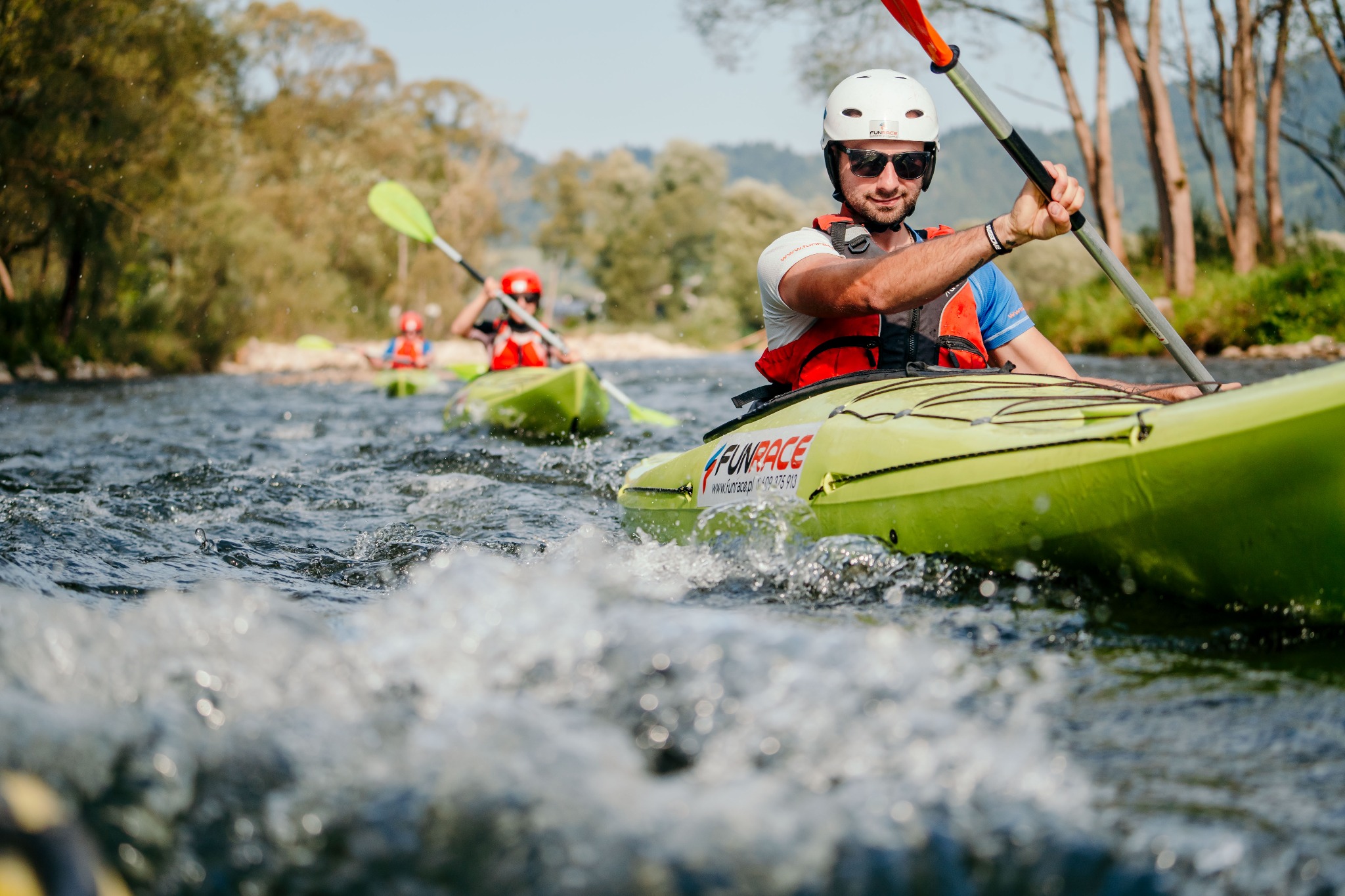 Spływ kajakowy po rzece z uczestnikami w kaskach i kamizelkach ratunkowych, widoczny mężczyzna z wiosłem w pierwszym planie i logo FUNRACE na kajaku.