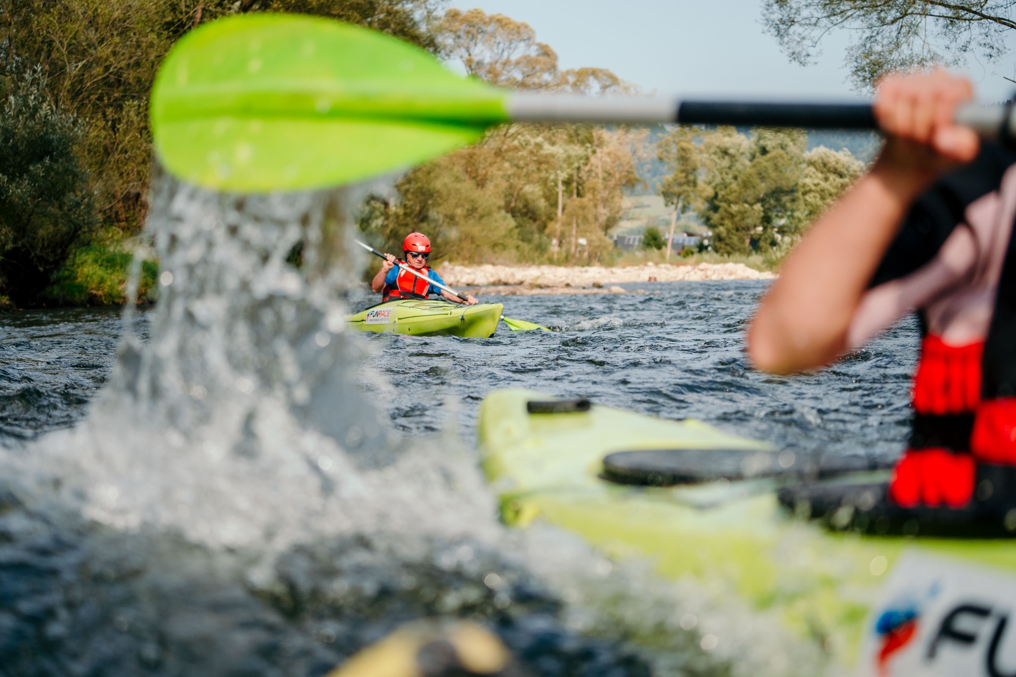 Kajakarze na rzece, wiosła w ruchu, woda pryska, w tle zalesiony brzeg.