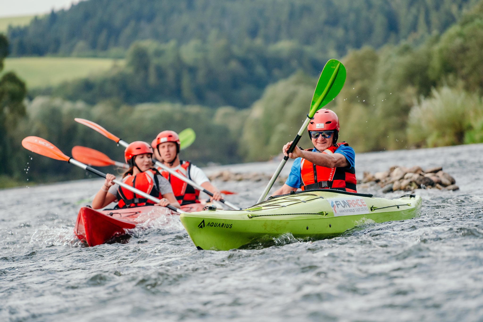 Grupa kajakarzy w kaskach i kamizelkach płynie po wzburzonej rzece, w tle zielone wzgórza, pierwszy plan to mężczyzna w zielonym kajaku z wiosłem w górze.