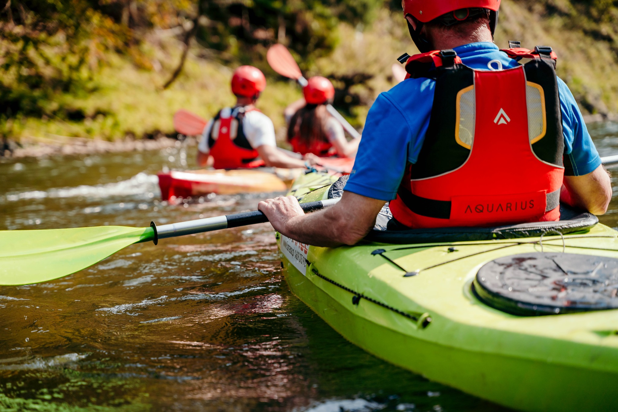 Osoby w kajakach płynące po rzece w słoneczny dzień, widok z perspektywy kajakarza trzymającego wiosło, w tle dwie osoby również w kajakach i czerwonych kaskach.