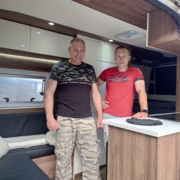 ARSO Meble Na Wymiar - Wnętrze kampera po przeróbce: dwaj mężczyźni prezentują zabudowę meblową z białymi szafkami, drewnianymi panelami i czarną tapicerką kanapy z puszystymi poduszkami.