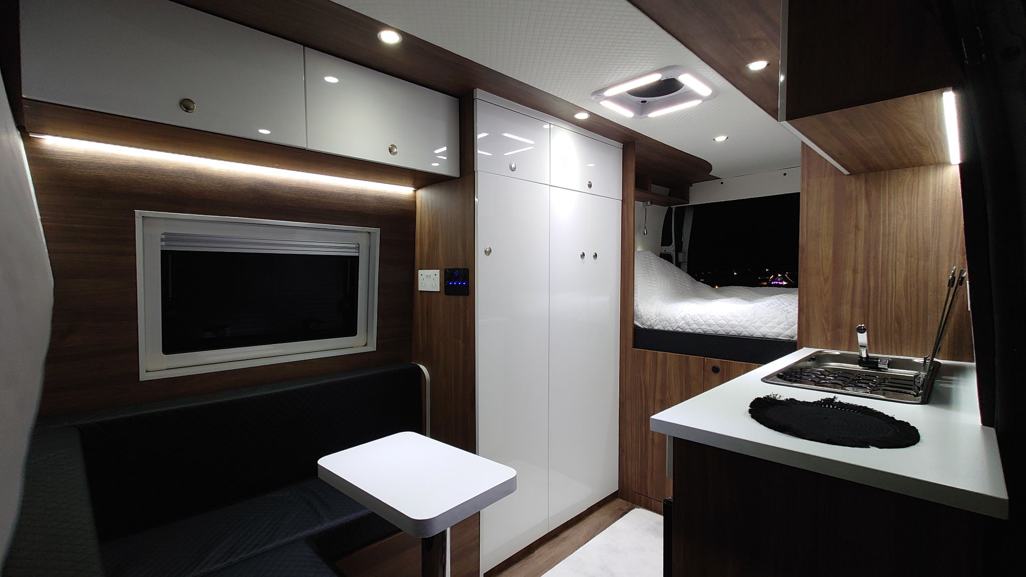Wnętrze kampera z zabudową na wymiar: białe szafy, drewniane panele, czarna kanapa, biały stolik, aneks kuchenny ze zlewem i widok na nocne miasto przez okno sypialni.