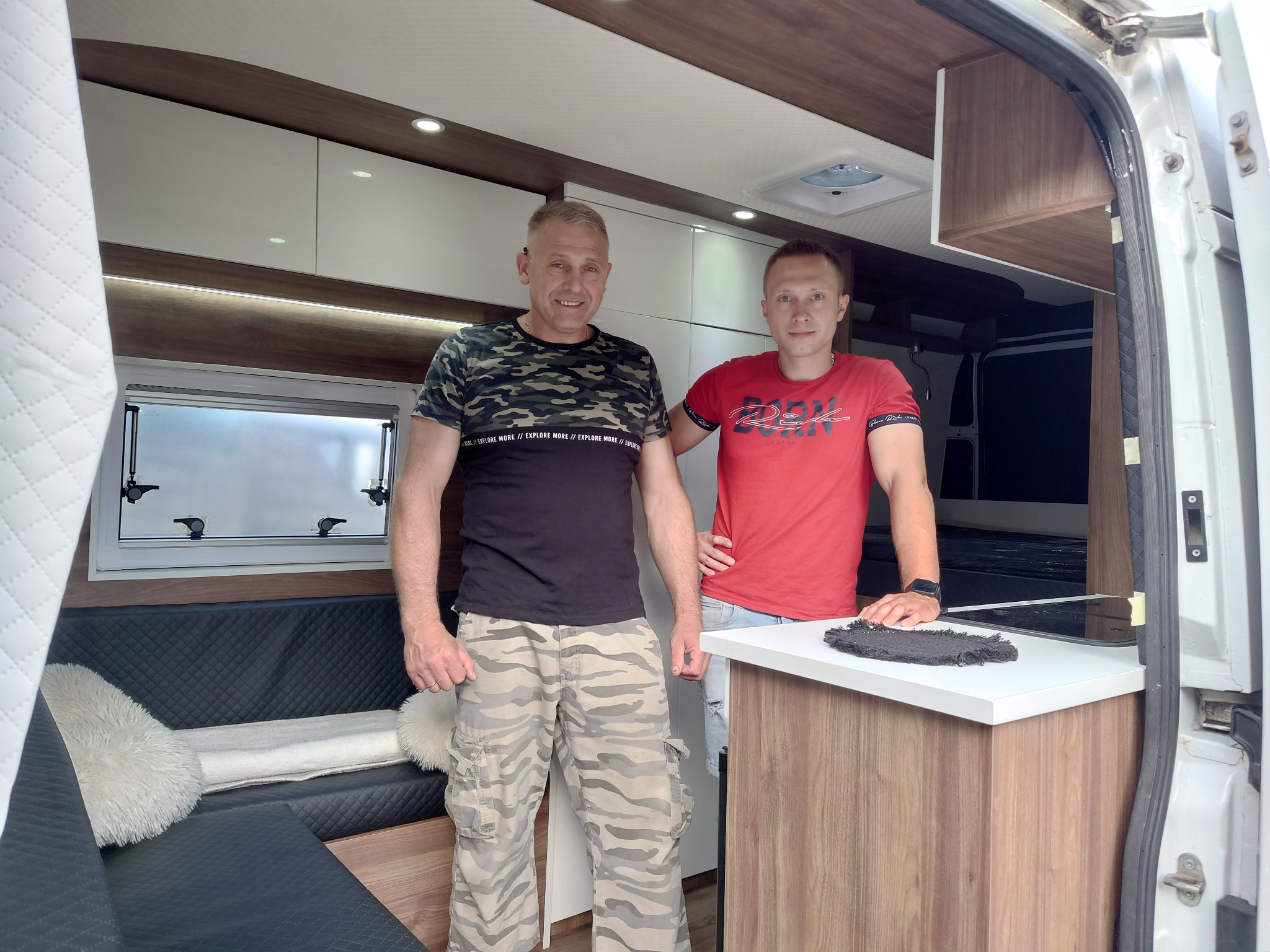 Wnętrze kampera po przeróbce: dwaj mężczyźni prezentują zabudowę meblową z białymi szafkami, drewnianymi panelami i czarną tapicerką kanapy z puszystymi poduszkami.
