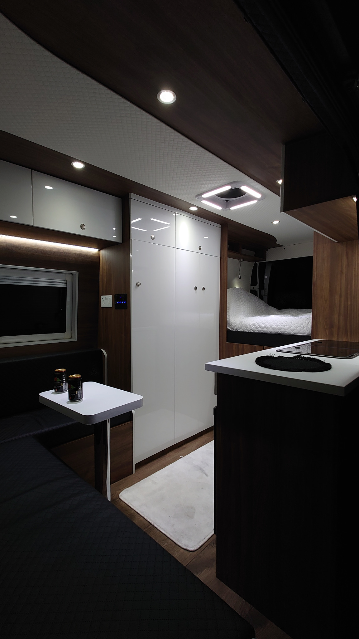 Wnętrze kampera z białą szafą wnękową, aneksem kuchennym i miejscem do spania, oświetlone punktowymi lampami sufitowymi.