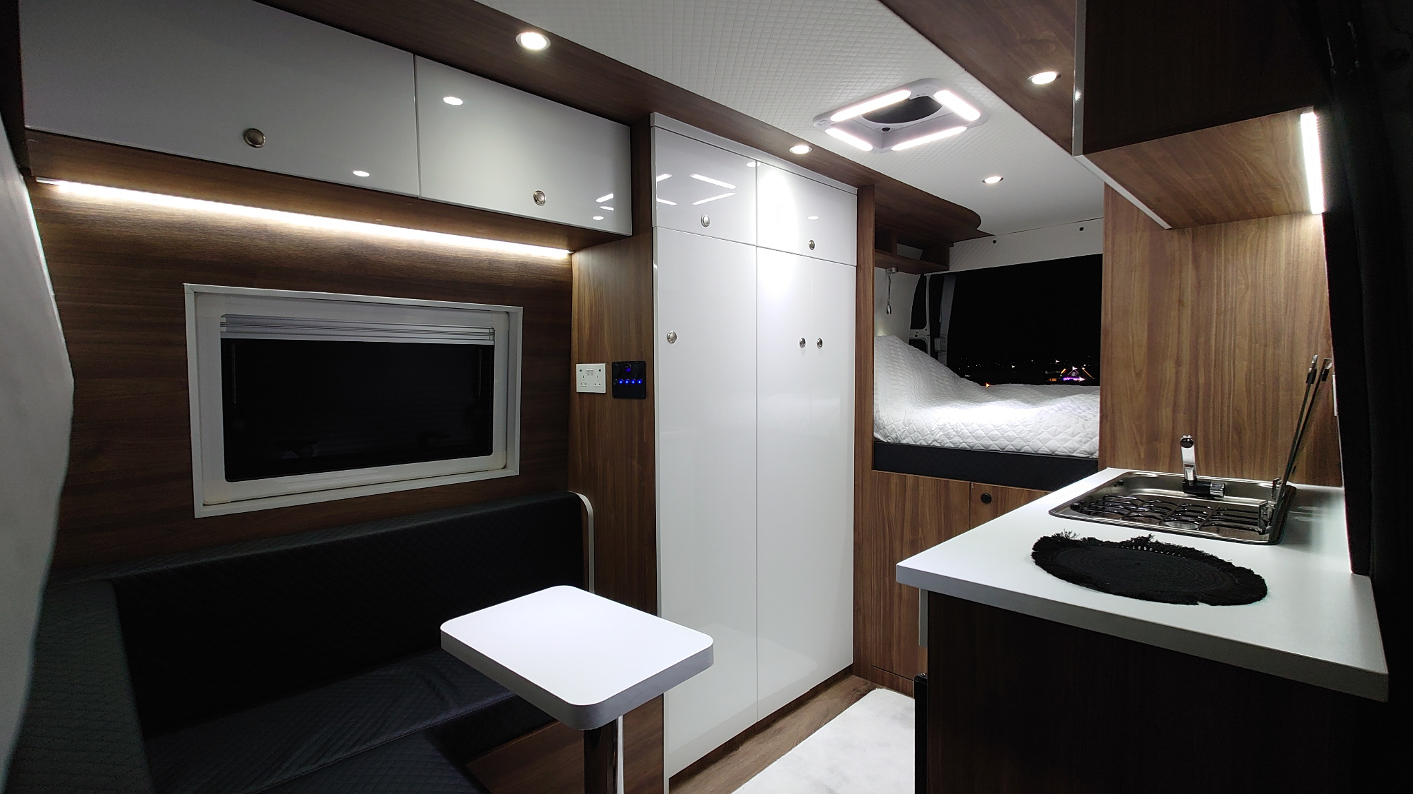 Wnętrze kampera z zabudową meblową: białe szafy, drewniane panele, czarna kanapa ze stolikiem, aneks kuchenny z metalowym zlewem i widok na łóżko z okna na nocny krajobraz.