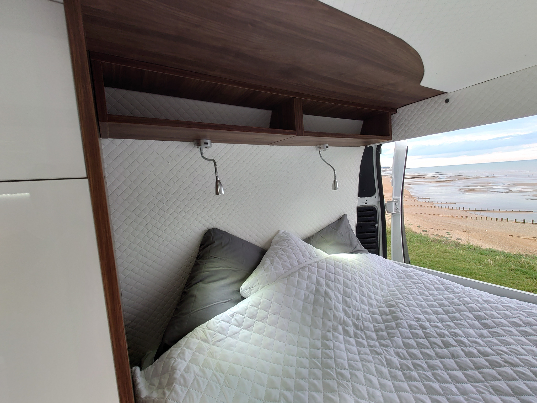 Wnętrze kampera z łóżkiem, szafką z ciemnego drewna i białym pikowanym zagłówkiem, widok na plażę przez otwarte drzwi.