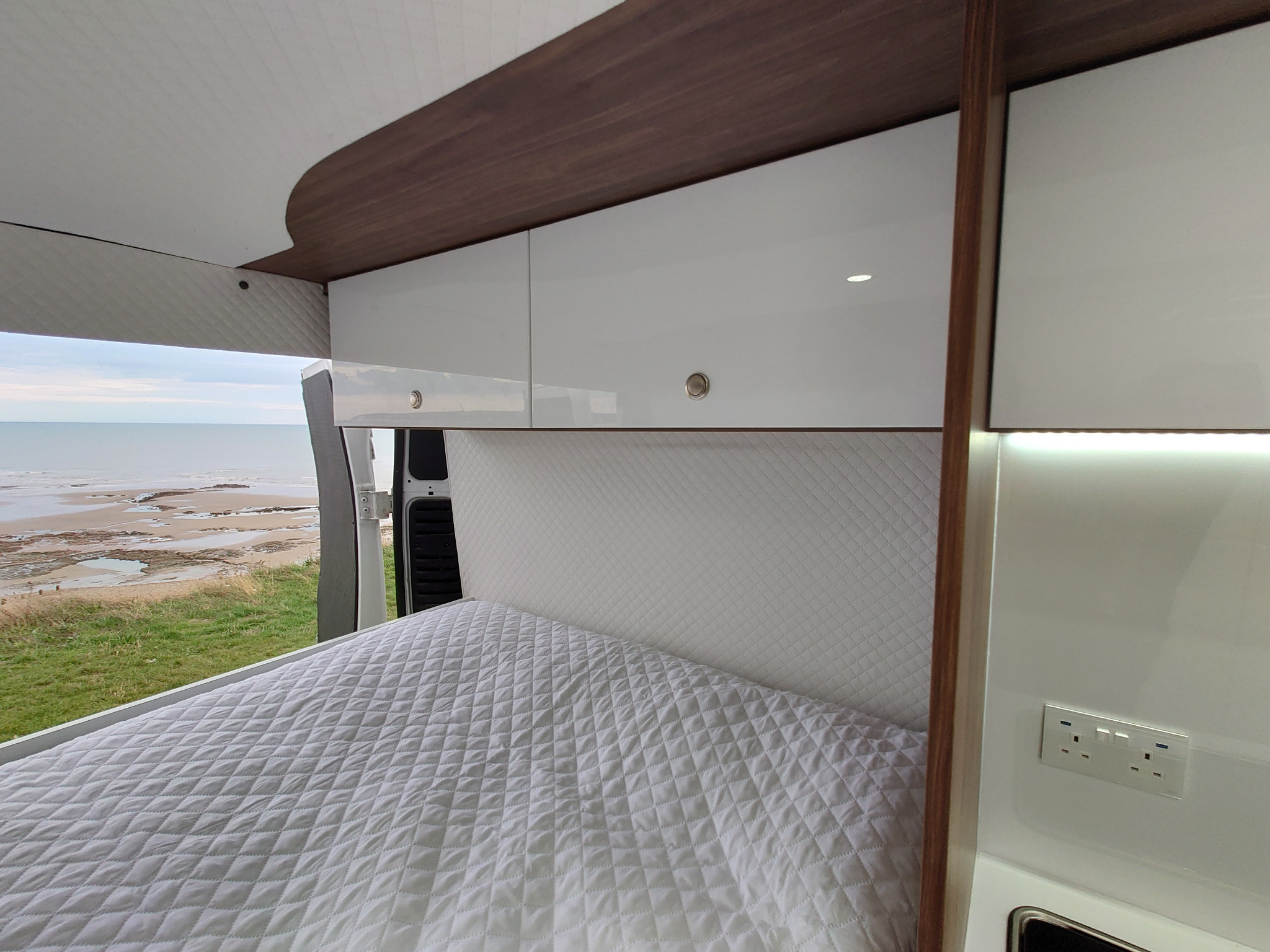 Wnętrze kampera z widokiem na plażę: białe szafki wiszące z drewnianym wykończeniem nad łóżkiem z pikowaną narzutą, gniazdko elektryczne na ścianie.