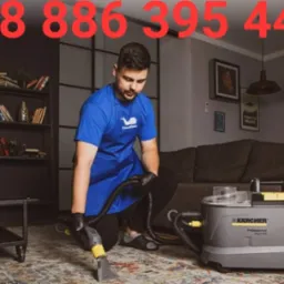 Mężczyzna w niebieskim fartuchu i czarnych rękawicach czyści dywan za pomocą odkurzacza piorącego Kärcher Professional Puzzi 400. W tle widoczny salon z szarą kanapą i regałem na książki.