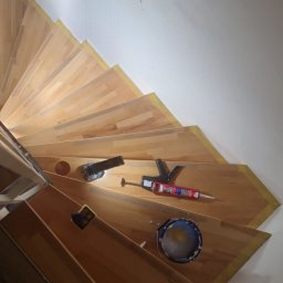 Radosław Kefer - Widok z góry na drewniane, kręte schody w trakcie renowacji, zabezpieczone taśmą malarską, z narzędziami (pistolet do silikonu, puszka farby) leżącymi na stopniach.