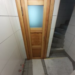 Radosław Kefer - Drewniane drzwi z szybą zamontowane w łazience wyłożonej białymi płytkami, widoczne zawory i fragment schodów w tle.