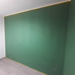 Radosław Kefer - Świeżo pomalowana ściana na zielono w pokoju, z żółtą taśmą malarską zabezpieczającą sufit i podłogę, widoczne gniazdka elektryczne.