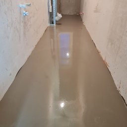 Radosław Kefer - Wylewka samopoziomująca w wąskim pomieszczeniu, widoczne ściany w stanie surowym i okno z widokiem na drzewa bez liści.