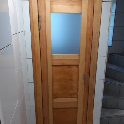 Radosław Kefer - Drewniane drzwi z przeszkleniem, zamontowane w łazience wyłożonej białymi, kwadratowymi płytkami, widoczne niedokończone prace wykończeniowe i wystające przewody.