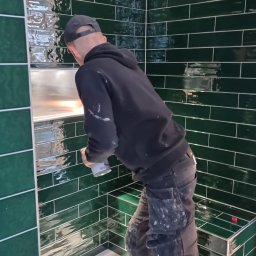 Radosław Kefer - Mężczyzna w czarnej bluzie i czapce, z plamami zaprawy na ubraniu, fuguje ciemnozielone, błyszczące płytki w łazience, widoczna wnęka w ścianie wyłożona płytkami, podłoga zabezpieczona folią.