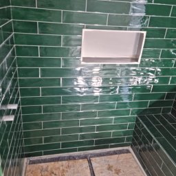 Radosław Kefer - Ściana prysznica wyłożona błyszczącymi, szmaragdowymi płytkami w układzie cegiełki z białymi fugami i wnęką na kosmetyki; widoczne niedokończone podłoże podłogowe.