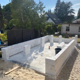 Radosław Kefer - Budowa ścian parteru z białych bloczków na fundamencie, widok z zewnątrz na plac budowy z czarnym wiadrem i piaskiem wokół, w tle dom jednorodzinny i zieleń.