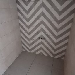 Radosław Kefer - Świeżo wykafelkowana, modernistyczna łazienka z szarymi płytkami ułożonymi we wzór zygzak na ścianie i podłodze, widoczne otwory na armaturę.