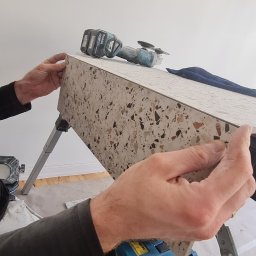 Radosław Kefer - Przygotowanie płytki lastryko na stojaku, szlifierka kątowa i niebieska skrzynka narzędziowa w tle, widoczne ręce glazurnika przytrzymujące płytkę.