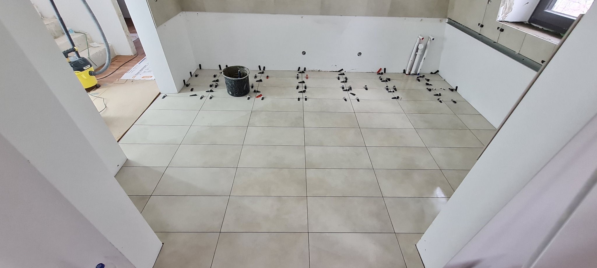 Jasne płytki podłogowe w trakcie układania, widoczne czarne kliny dystansowe, wiadro i odkurzacz przemysłowy w tle.