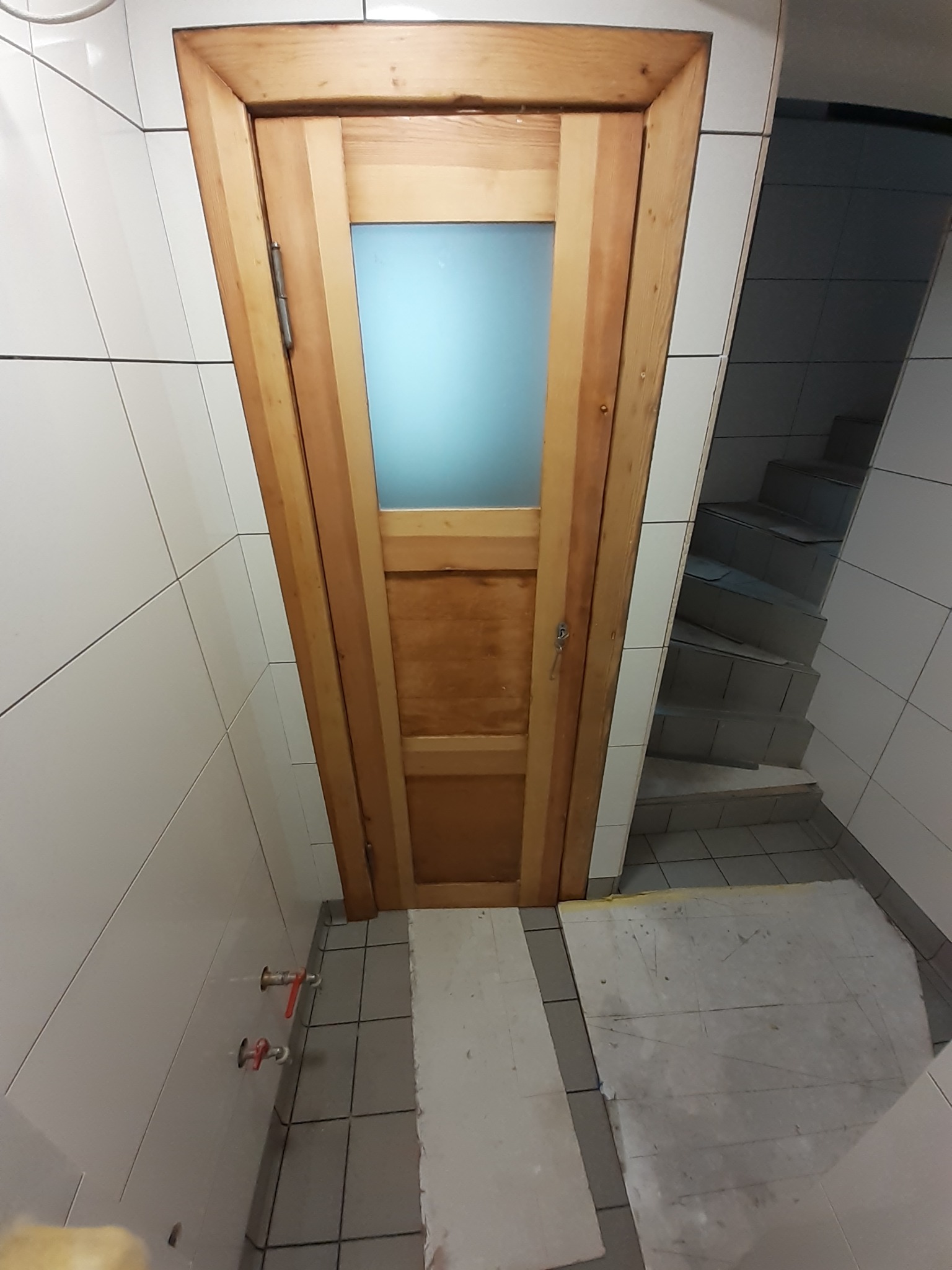 Drewniane drzwi z szybą zamontowane w łazience wyłożonej białymi płytkami, widoczne zawory i fragment schodów w tle.