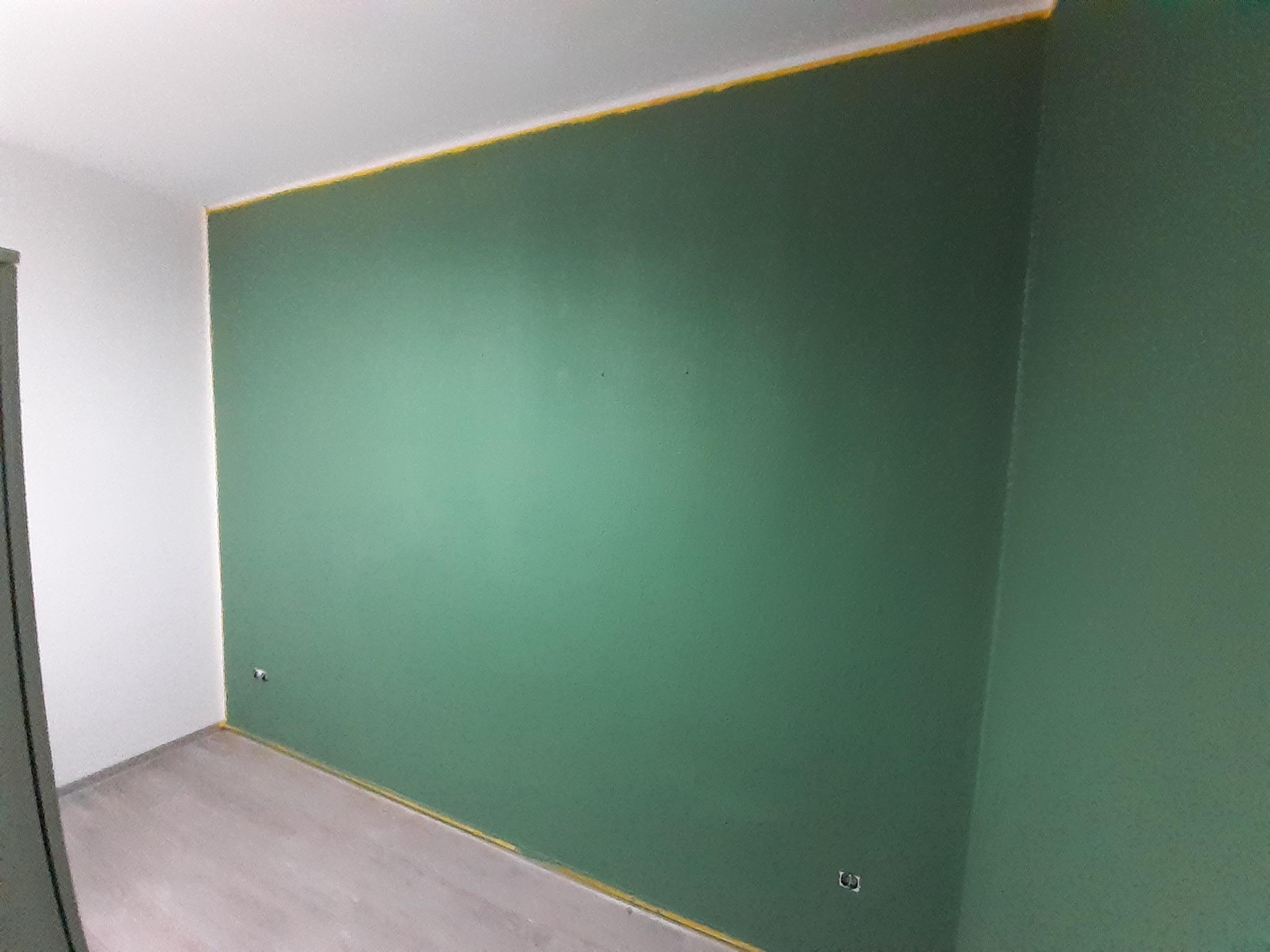 Świeżo pomalowana ściana na zielono w pokoju, z żółtą taśmą malarską zabezpieczającą sufit i podłogę, widoczne gniazdka elektryczne.