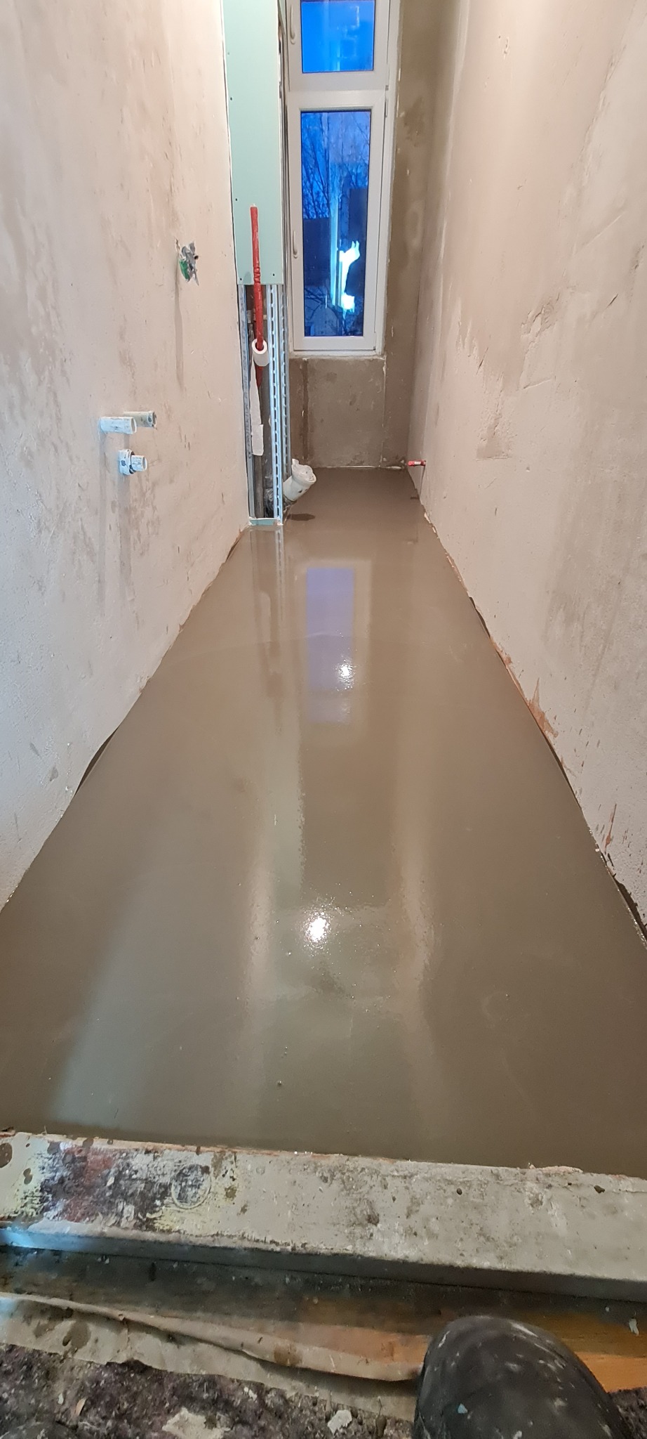 Wylewka samopoziomująca w wąskim pomieszczeniu, widoczne ściany w stanie surowym i okno z widokiem na drzewa bez liści.