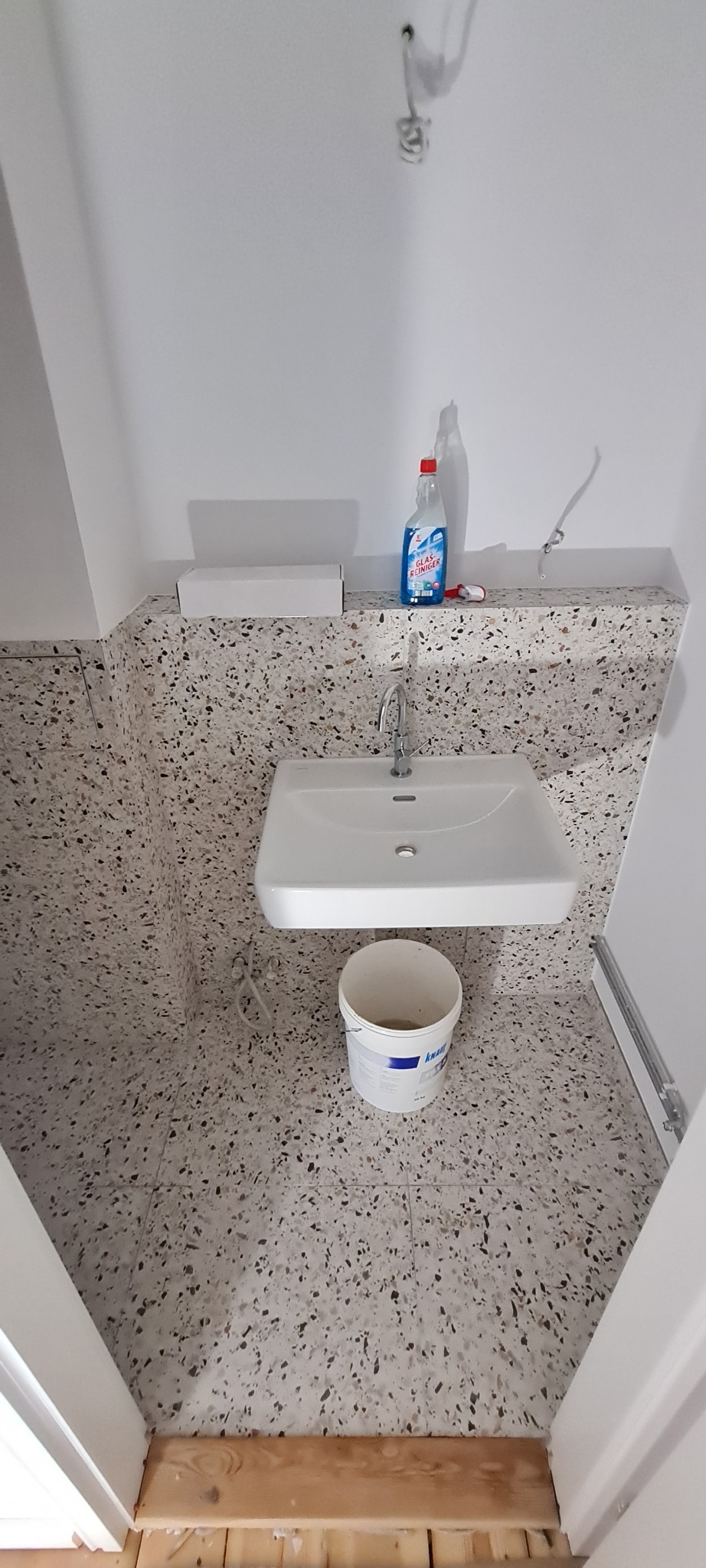 Łazienka w trakcie remontu: biała umywalka zamontowana na ścianie z terazzo, wiadro z zaprawą obok, widoczne instalacje, surowy stan wykończenia podłogi przy drzwiach.