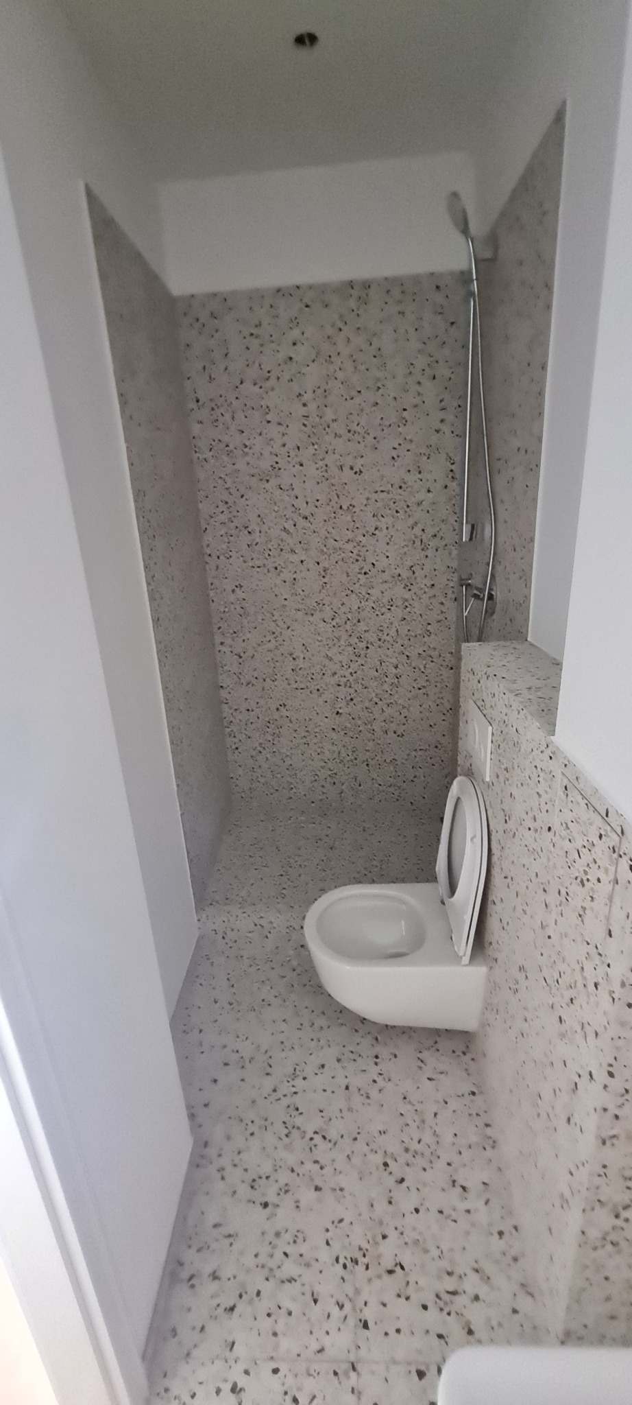 Nowoczesna łazienka z toaletą podwieszaną i prysznicem, wykończona płytkami imitującymi lastryko na ścianach i podłodze, z minimalistyczną armaturą.