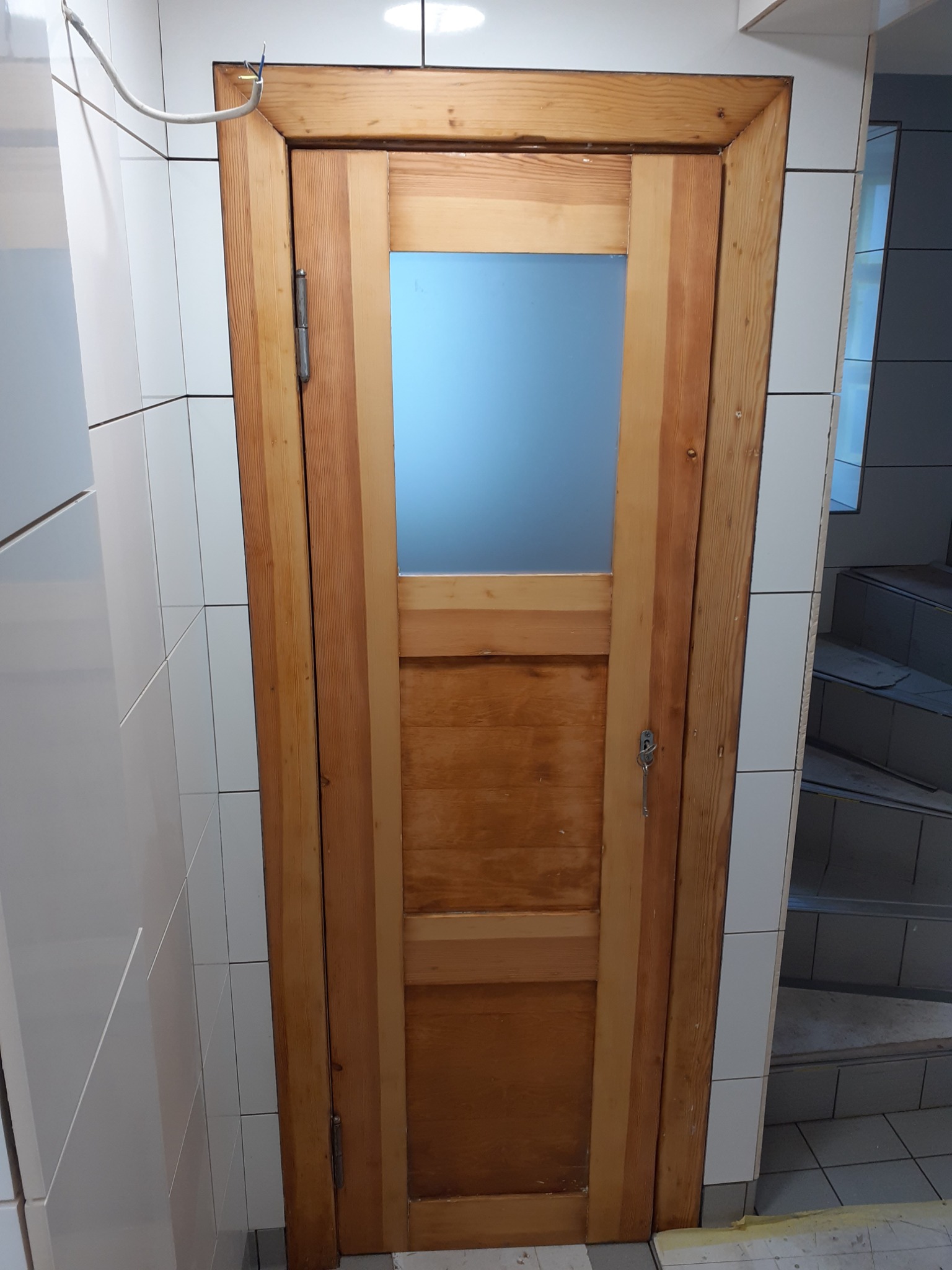 Drewniane drzwi z przeszkleniem, zamontowane w łazience wyłożonej białymi, kwadratowymi płytkami, widoczne niedokończone prace wykończeniowe i wystające przewody.