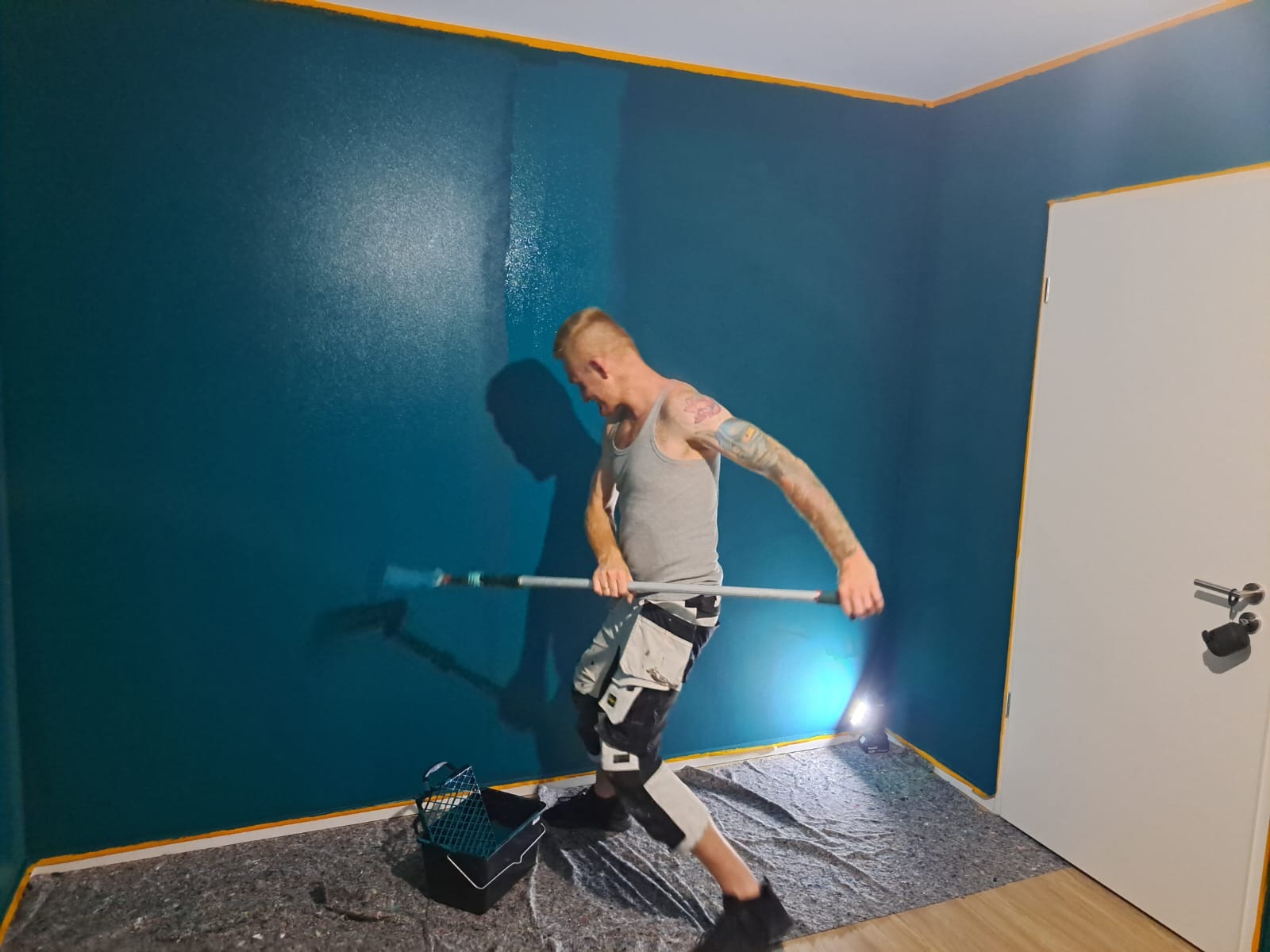 Mężczyzna z tatuażami maluje ścianę na ciemnozielono za pomocą wałka na długim kiju, widoczny kuweta z farbą, podłoga zabezpieczona folią malarską, ściana częściowo pomalowana, widoczne taśmy...