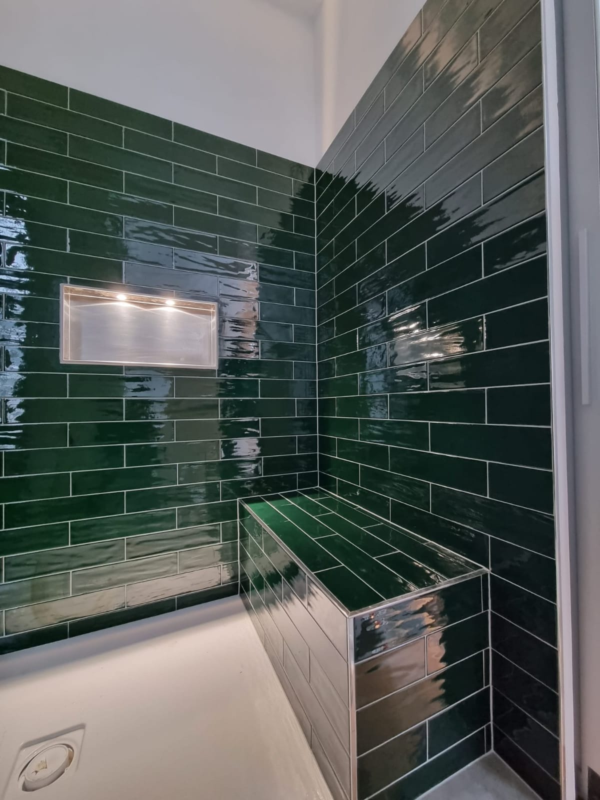 Wykończona łazienka z ciemnozielonymi, błyszczącymi płytkami ułożonymi horyzontalnie, wbudowana półka z oświetleniem i siedzisko z płytek.