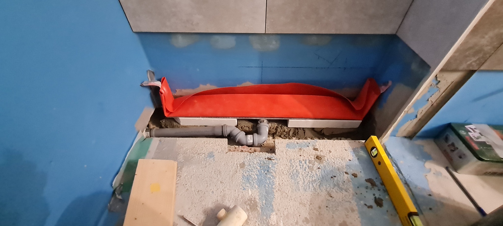 Przygotowanie odpływu liniowego w łazience: widoczna hydroizolacja w kolorze pomarańczowym, rury kanalizacyjne, poziomica i fragmenty płytek na ścianie, etap prac przed położeniem płytek.