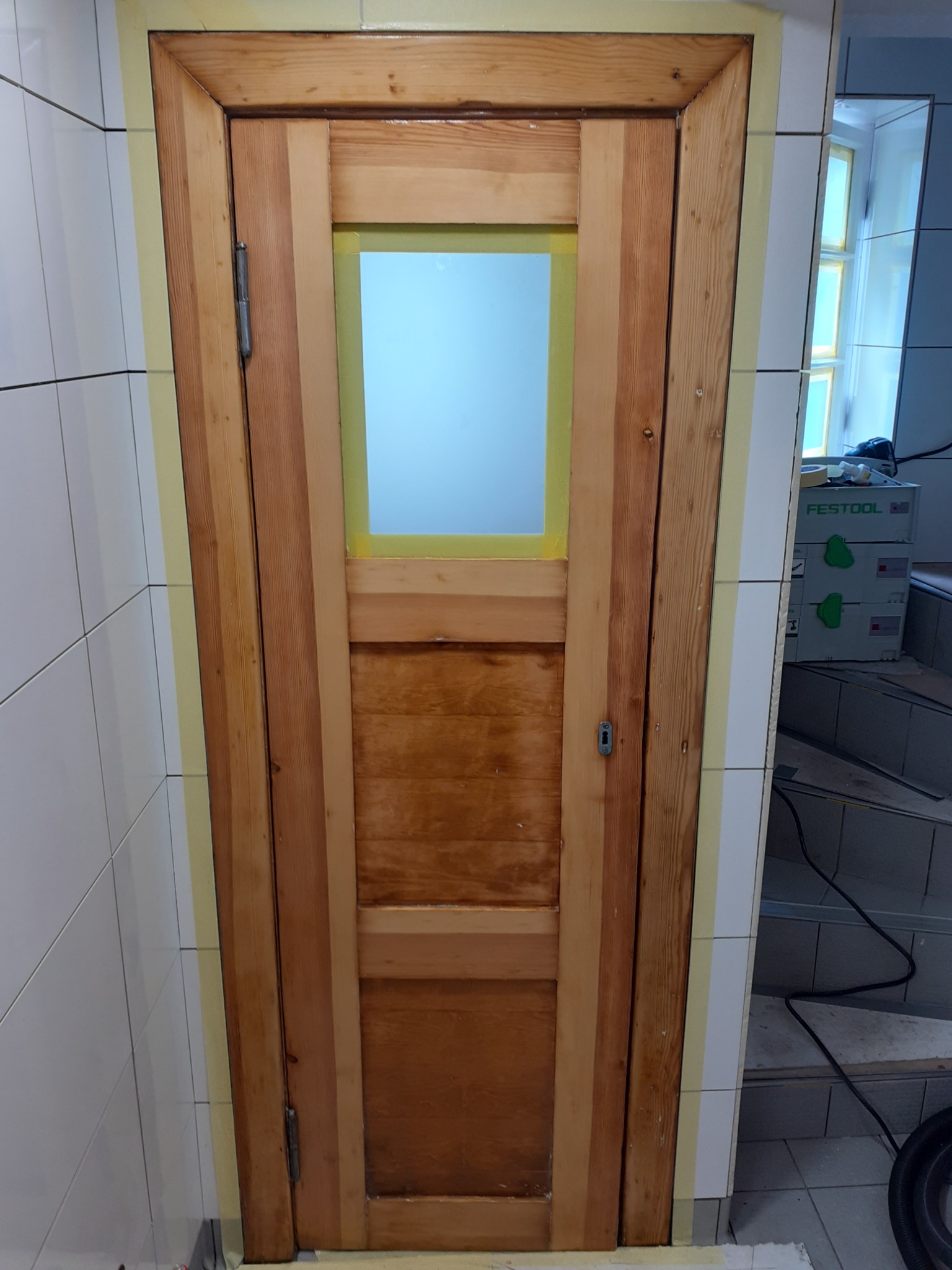 Drewniane drzwi z szybą, zabezpieczone taśmą malarską na styku z białą, kwadratową glazurą, w tle widoczny sprzęt Festool.