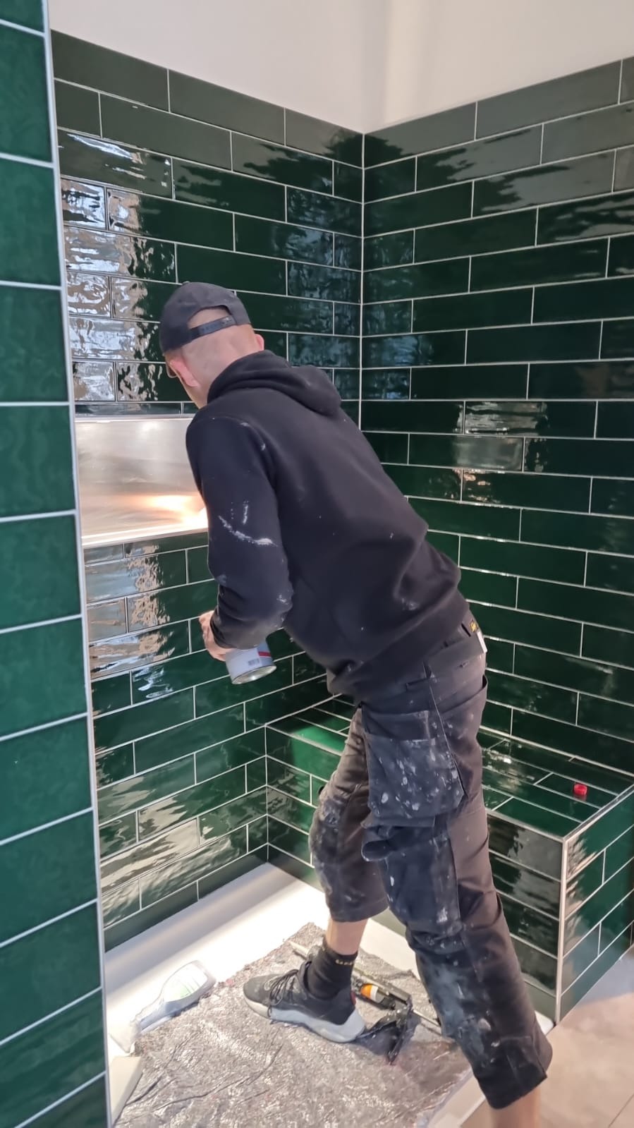 Mężczyzna w czarnej bluzie i czapce, z plamami zaprawy na ubraniu, fuguje ciemnozielone, błyszczące płytki w łazience, widoczna wnęka w ścianie wyłożona płytkami, podłoga zabezpieczona folią.