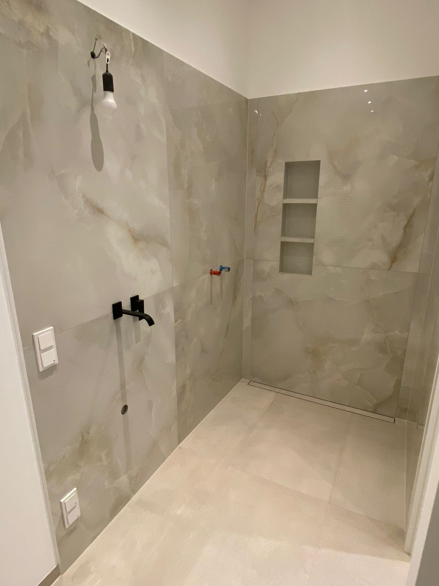 Nowoczesna kabina prysznicowa z beżowymi płytkami imitującymi marmur, czarną armaturą i odpływem liniowym, oświetlona wiszącą żarówką na kablu.