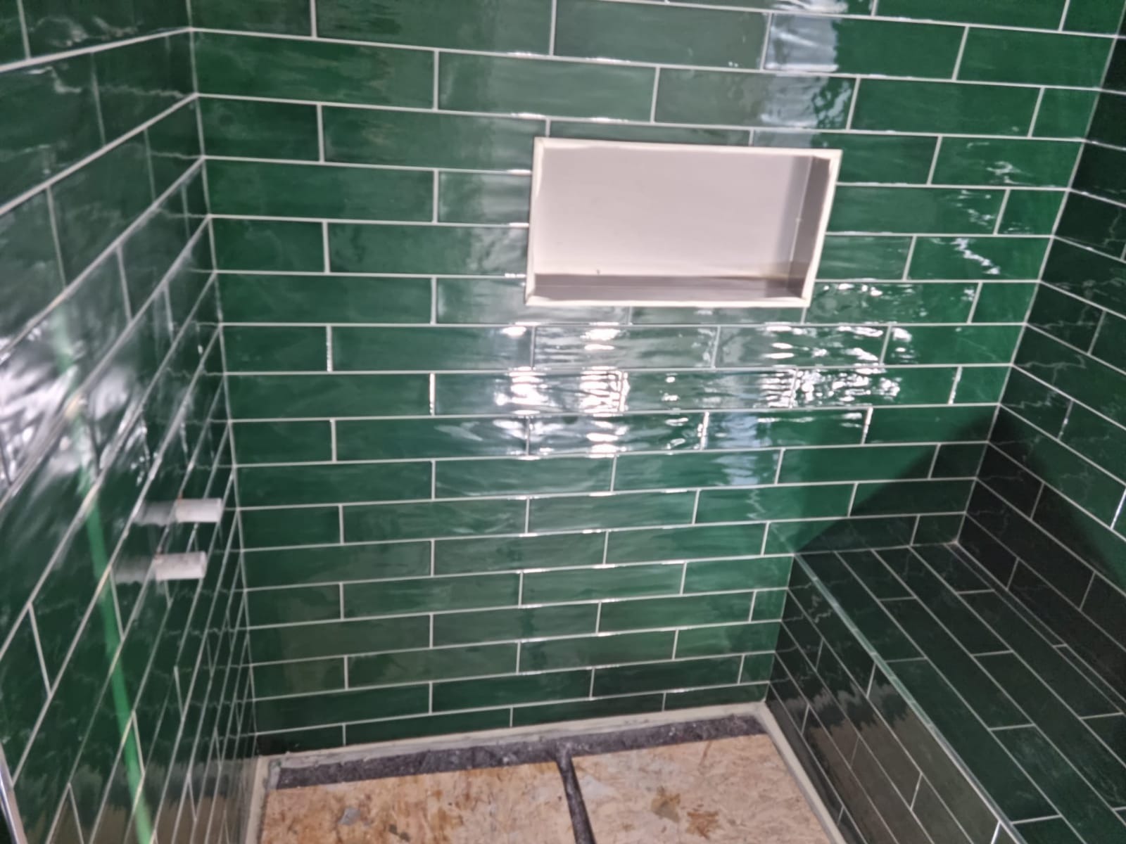 Ściana prysznica wyłożona błyszczącymi, szmaragdowymi płytkami w układzie cegiełki z białymi fugami i wnęką na kosmetyki; widoczne niedokończone podłoże podłogowe.