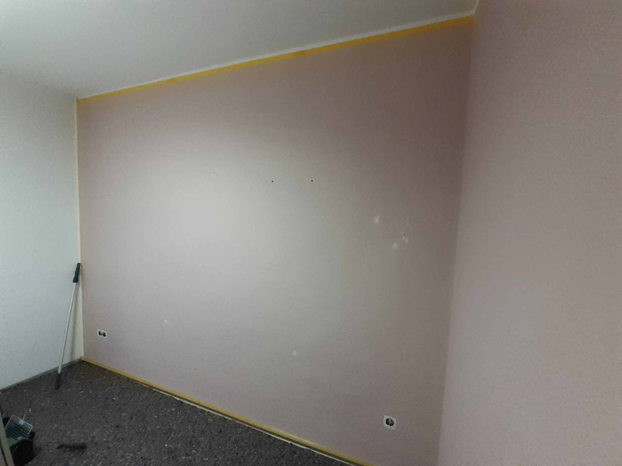 Ściana w pokoju przygotowana do malowania z taśmą malarską na górze i dole, widoczne gniazdka elektryczne i wałek malarski oparty o ścianę.