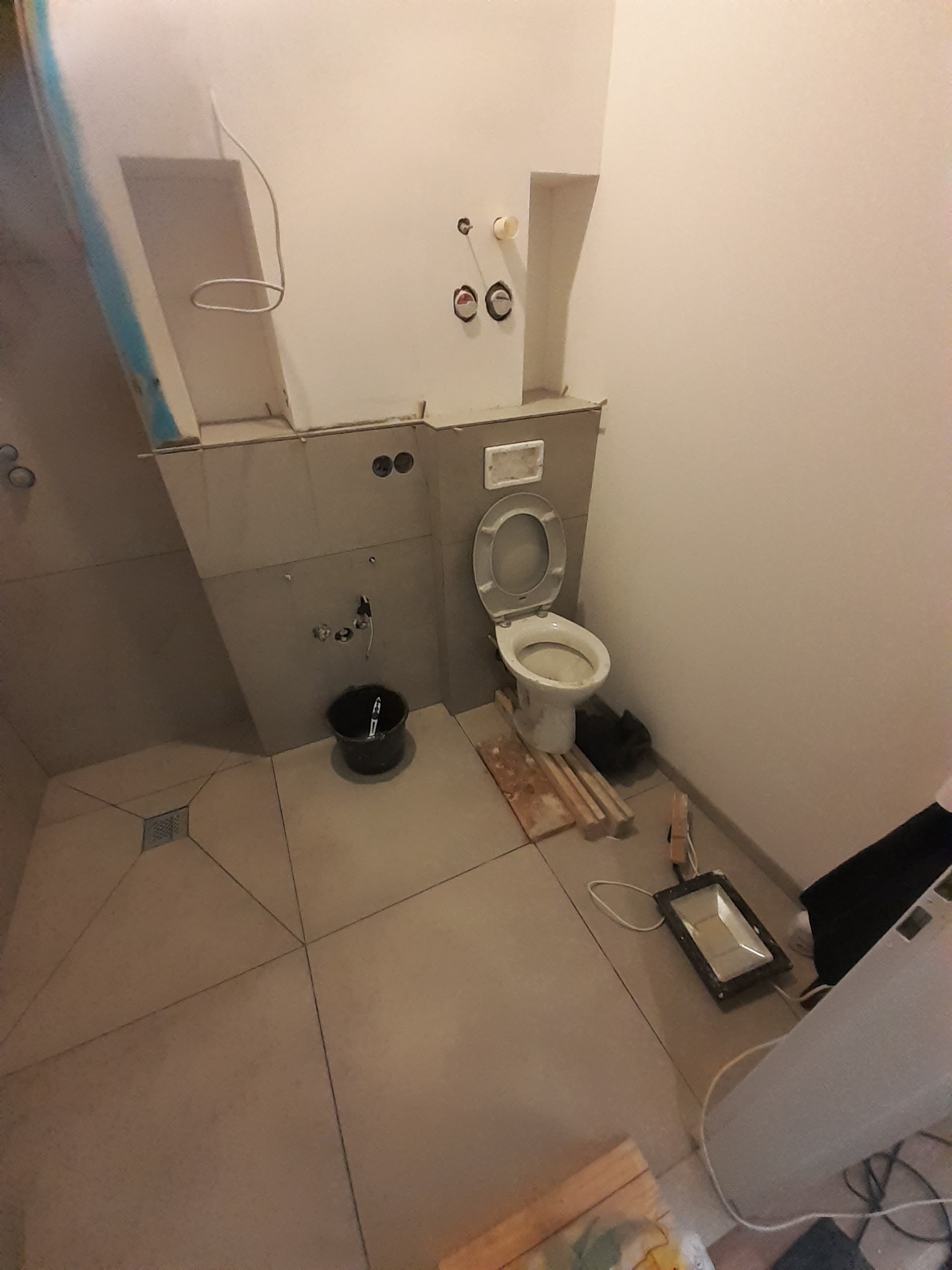 Remontowana łazienka w Sycowie: widok na częściowo wykończoną kabinę prysznicową z odpływem w podłodze, podwieszaną toaletę w trakcie montażu, wiadro i lampę budowlaną.