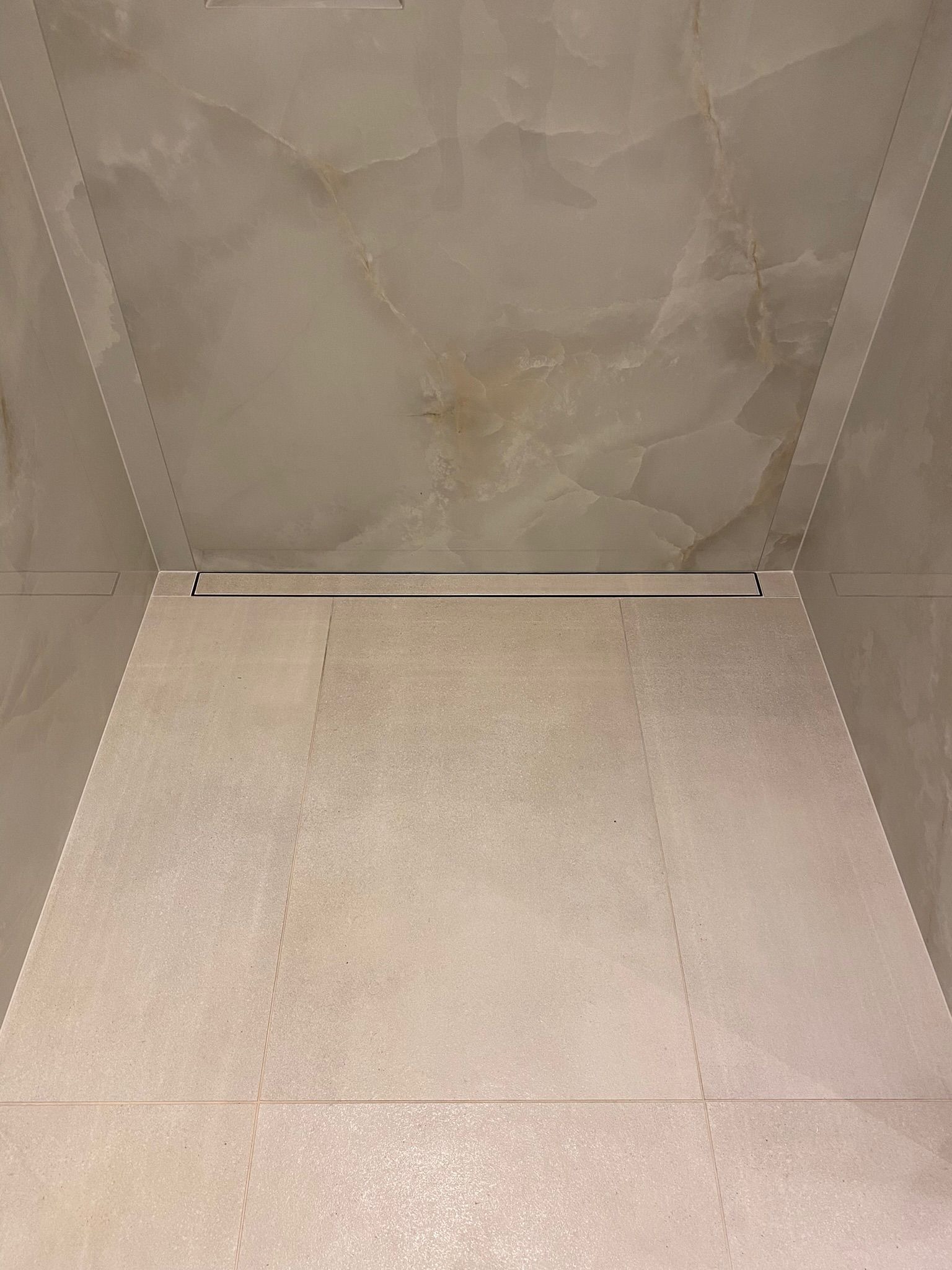 Beżowe płytki podłogowe i marmuropodobne płytki ścienne w kabinie prysznicowej z odpływem liniowym.