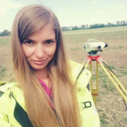 Kobieta w kurtce ostrzegawczej robiąca selfie na tle pola z niwelatorem optycznym na statywie.