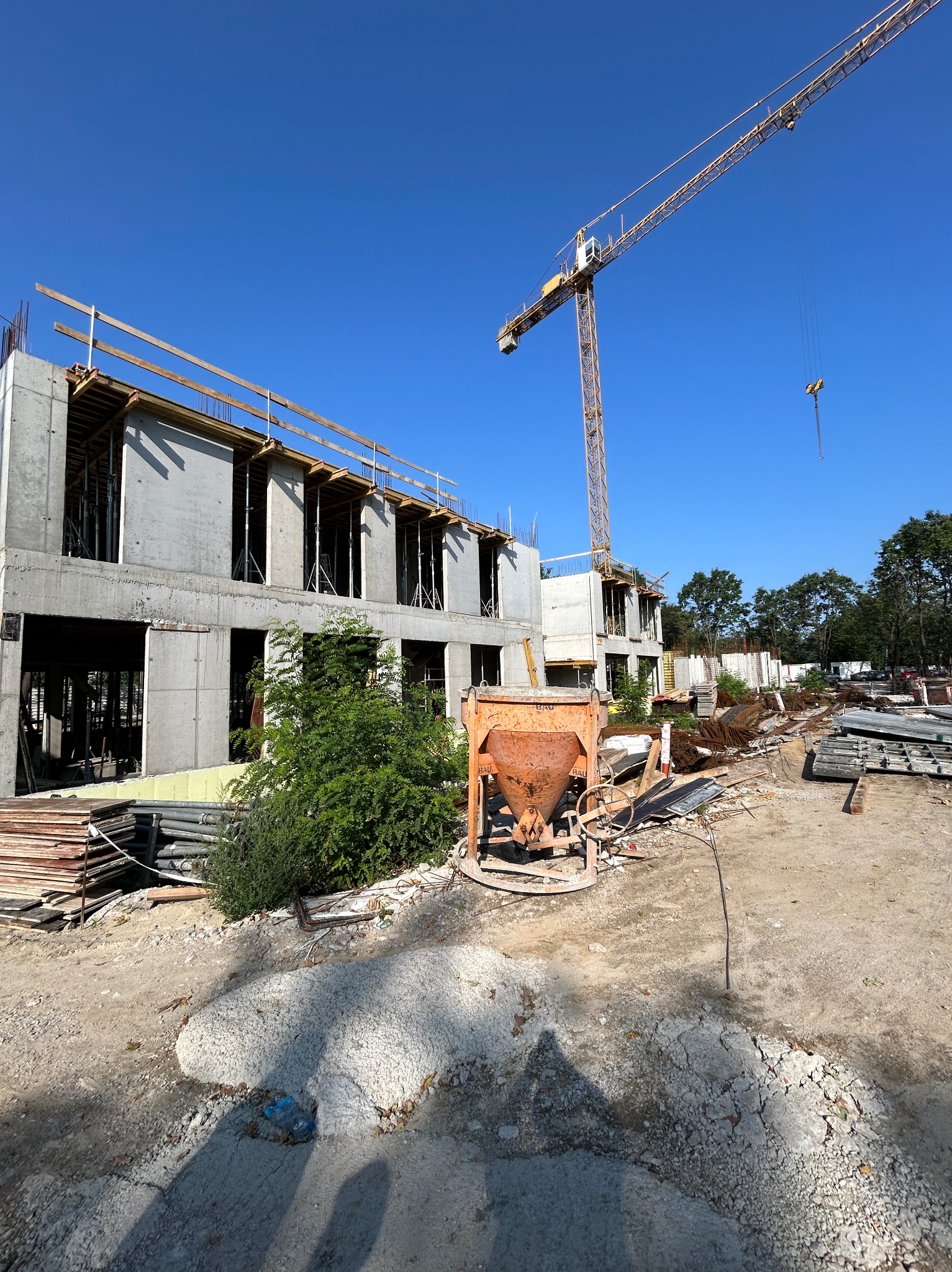 Budowa dwukondygnacyjnego budynku w stanie surowym otwartym, z żurawiem budowlanym i pomarańczową betoniarką na pierwszym planie w słoneczny dzień.