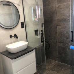 FIXARS - Nowoczesna łazienka z kabiną prysznicową typu walk-in, umywalką nablatową i okrągłym lustrem w czarnej ramie. Dominują szarości i biel, podkreślone czarnymi akcentami.