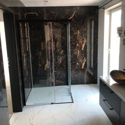 FIXARS - Elegancka łazienka z czarnymi płytkami imitującymi marmur i kabiną prysznicową ze złotymi akcentami. Biała marmurowa podłoga i czarna szafka pod umywalką.