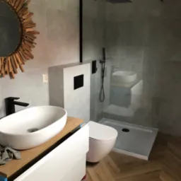 Nowoczesna łazienka z kabiną typu walk-in, umywalką nablatową na drewnianym blacie i lustrem w drewnianej ramie. Na podłodze panele ułożone w jodełkę.