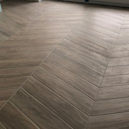 FIXARS - Ukończona podłoga z płytek imitujących drewno, ułożonych w jodełkę, przy oknie zabezpieczonym taśmą malarską. Ciemniejsze fugi podkreślają wzór.