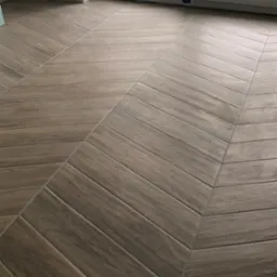 Ukończona podłoga z płytek imitujących drewno, ułożonych w jodełkę, przy oknie zabezpieczonym taśmą malarską. Ciemniejsze fugi podkreślają wzór.