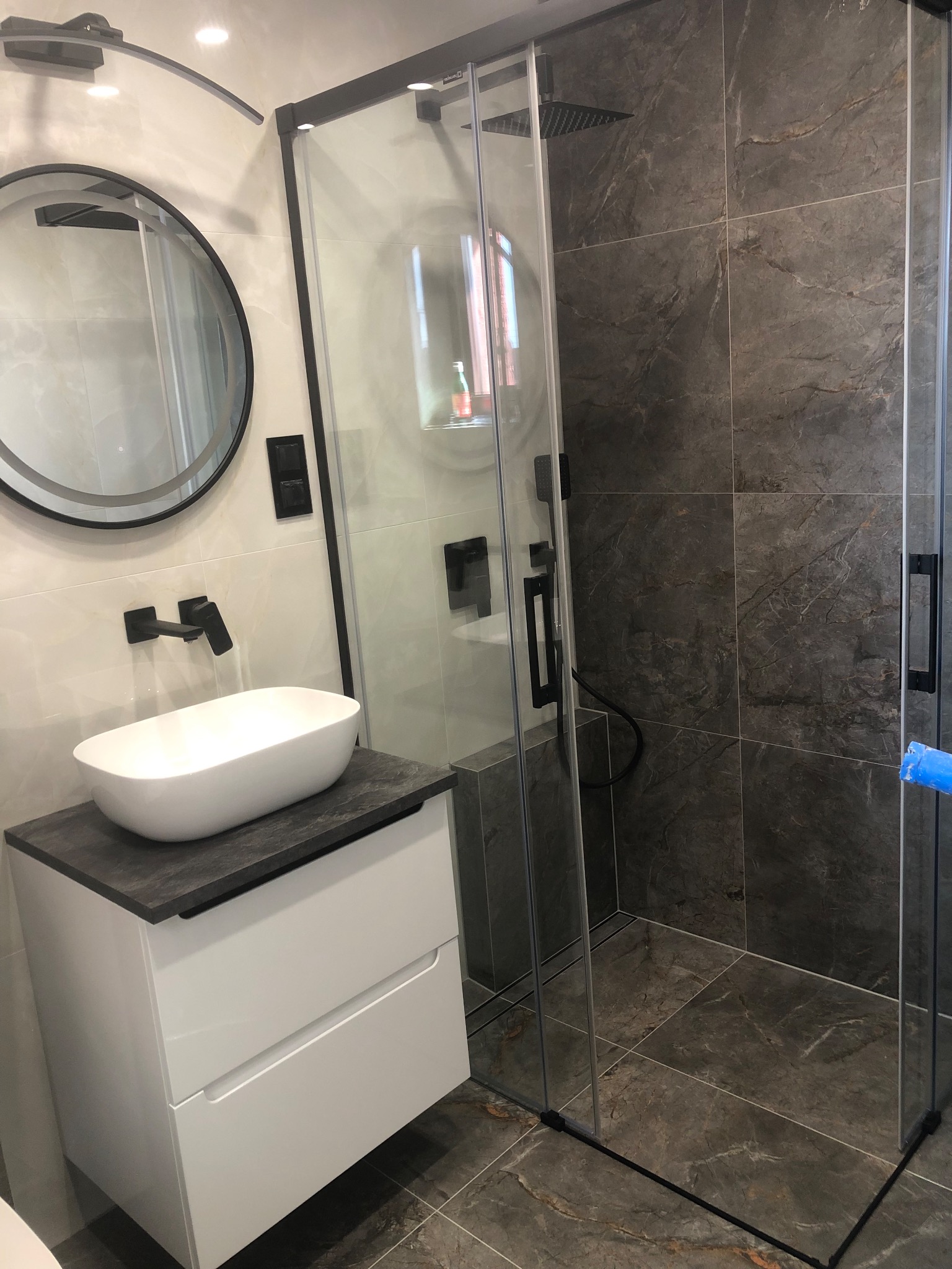 Nowoczesna łazienka z kabiną prysznicową typu walk-in, umywalką nablatową i okrągłym lustrem w czarnej ramie. Dominują szarości i biel, podkreślone czarnymi akcentami.