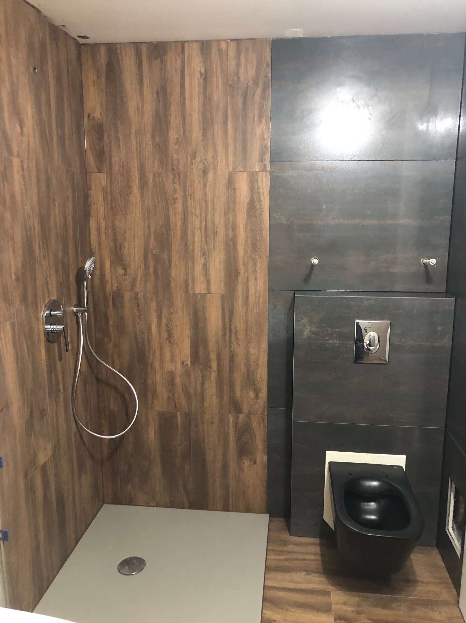 Nowoczesna łazienka z drewnopodobnymi i grafitowymi płytkami, kabiną prysznicową typu walk-in i podwieszaną toaletą. Chromowana armatura podkreśla minimalistyczny design.