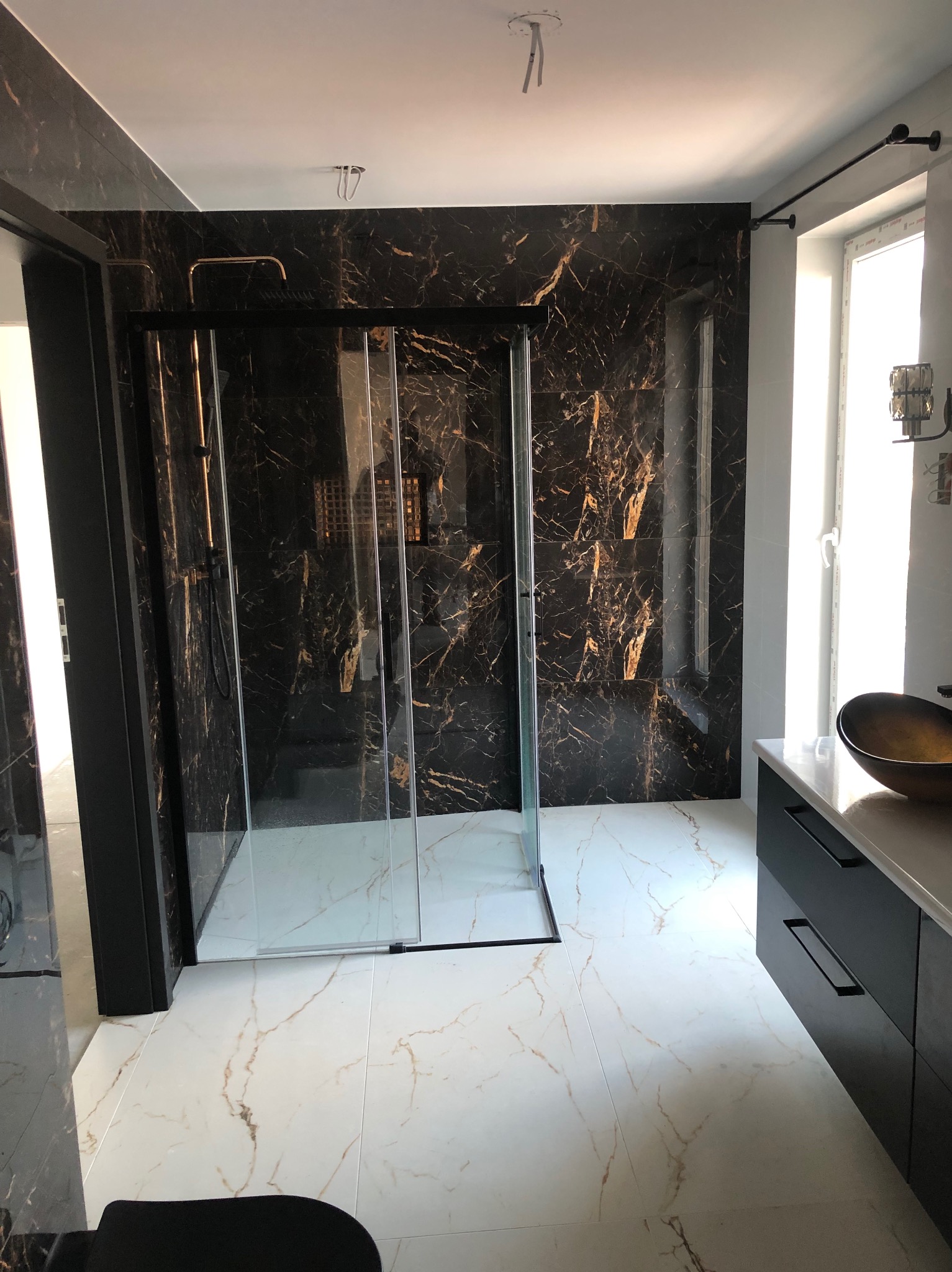 Elegancka łazienka z czarnymi płytkami imitującymi marmur i kabiną prysznicową ze złotymi akcentami. Biała marmurowa podłoga i czarna szafka pod umywalką.