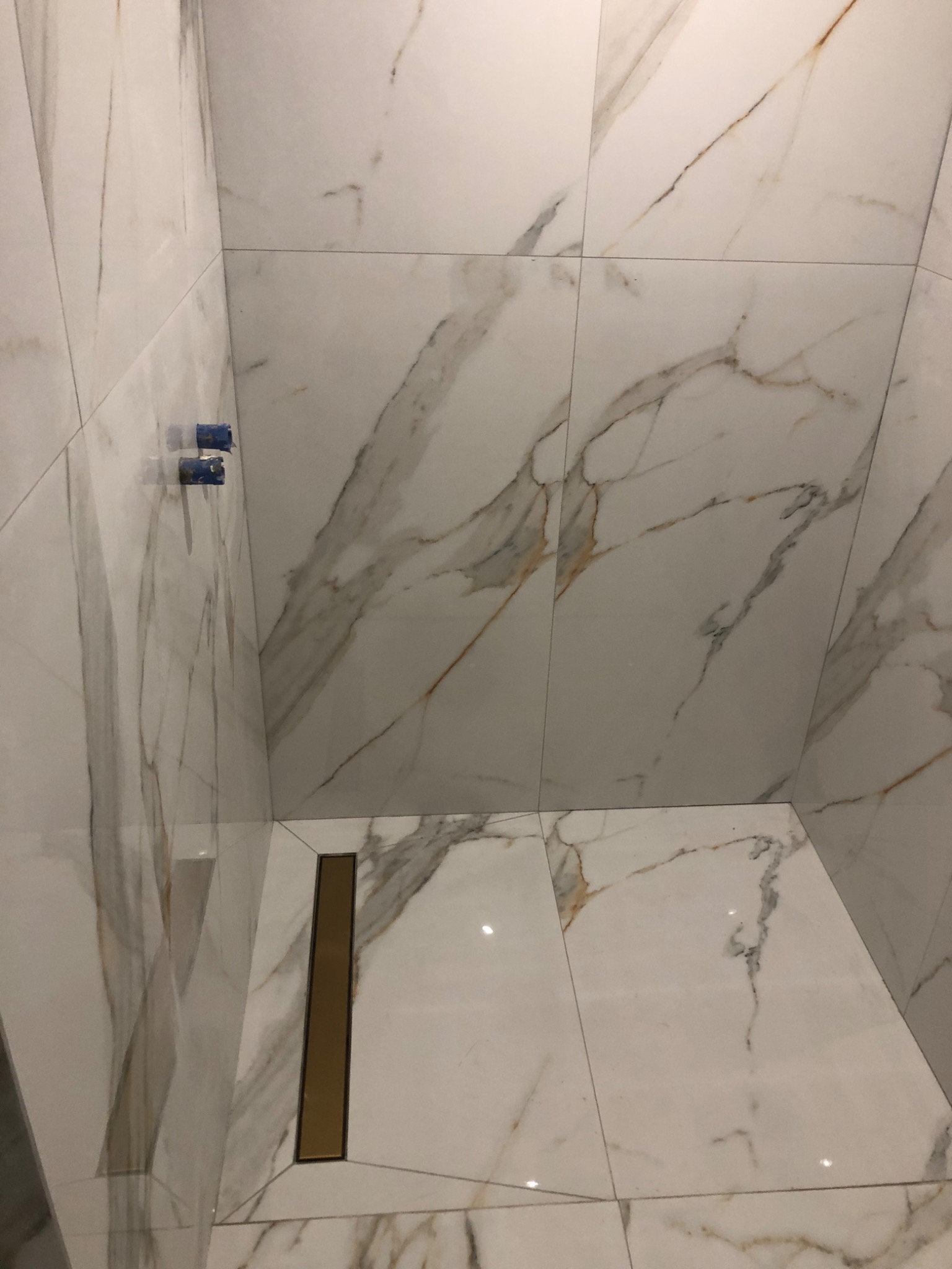 Wykończona kabina prysznicowa z białymi płytkami imitującymi marmur i złotym odpływem liniowym. Widoczne niebieskie zaślepki na rurach.